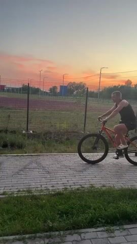 A man is riding a bright orange bicycle on a paved path. He is wearing a black tank top and patterned shorts. The bicycle has thick tires, typical of a mountain bike. The man is looking back towards the camera with a slight smile. The setting appears to be an outdoor recreational area, possibly a park or sports complex. In the background, there is a fence, a grassy field, and several multi-story buildings. The sky is a soft gradient of orange and pink, indicating that it is either sunrise or sunset. There are streetlights visible in the distance. The grass along the path is green and appears well-maintained. No visible text is present in the media. The overall mood is relaxed and pleasant, suggesting leisure activity during a beautiful time of day.