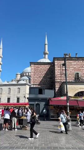 The video pans across a bustling outdoor plaza under a clear blue sky. In the background, a large, ornate mosque with two tall minarets dominates the scene. The mosque features a central dome and is constructed with light-colored stone. Several people are walking through the plaza, some in groups, others individually. In the foreground, a woman with dark hair walks towards the camera, carrying a black handbag and wearing black pants and white sneakers. To her left, a vendor cart with a red canopy is visible, displaying various items. A couple of other vendor carts are also present. Some people are browsing at the stalls. The ground is paved with interlocking stones. The overall atmosphere appears to be lively and tourist-oriented, likely in a city with significant historical and architectural landmarks.