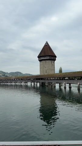 The video shows a body of water with a long, covered bridge spanning across it. At one end of the bridge is a tall stone tower with a brown, conical roof. The tower's reflection is visible in the water. On the far side of the water, there's a row of buildings with varied architectural styles. Some have spires or turrets, and others appear to be residential or commercial. The sky is overcast, and the overall lighting is muted. The camera pans to reveal the buildings further down the shoreline. The water is calm, with slight ripples, and there's a railing in the foreground of the shot.