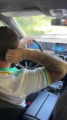 A man is driving a Mercedes-Benz. The man has dark hair, a beard, sunglasses, a white polo shirt with green and yellow trim, and tattoos on his left arm. He is looking ahead at the road. The car has a modern interior with a digital dashboard and a phone sitting in the center console. The view out of the windshield shows a sunny day with trees lining the road. The video is shot from the backseat of the car.