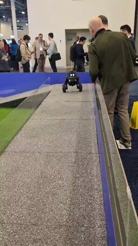 The video shows a small black robot with illuminated green eyes moving forward on a gray, carpeted ramp, surrounded by a blue and clear barrier.  A bald man wearing a green jacket and tan pants stands close to the barrier, watching the robot. He is facing the robot with his hands lightly touching the barrier. Other people are visible in the background, walking and standing around what appears to be a convention or trade show. A yellow bag is visible to the right of the man, and a small patch of artificial green grass is seen on the left side of the ramp.