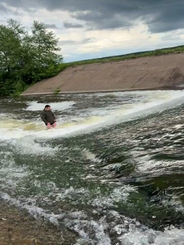 A man is standing in the middle of a waterfall in a river. He's wearing shorts and a long-sleeved shirt. The water is rushing around him, and there's a man-made dam behind him. On the left, there are a few trees and brush. The sky is overcast.
