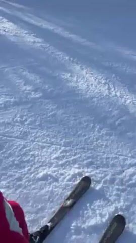 A person is skiing downhill on a sunny day. The person is wearing a red hooded jacket. The snow is white with some tracks and shadows. The trees are visible in the background. The camera is positioned from the perspective of the skier.