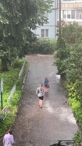 This video captures a rainy day scene in Tver, Russia. The primary subjects are three individuals walking on a wet, paved pathway. The path is bordered by lush green vegetation and a white metal fence on the left, and dense foliage on the right. In the background, a multi-story apartment building with light blue siding and windows is visible.

The event taking place is people walking during a heavy downpour. The rain is a prominent feature, creating puddles on the pathway and splashing around the walkers. One person in the foreground, wearing a light purple top and black shorts, is walking away from the camera. Further up the path, two individuals are walking in the same direction; one in a grey top and black shorts, and another, partially obscured, in a red top. They appear to be walking quickly, perhaps trying to find shelter.

The weather is clearly rainy, with visible streaks of water falling from the sky. The time of day is not explicitly indicated, but the lighting suggests it could be daytime, possibly overcast due to the rain. The setting is urban, likely a residential area given the apartment building and pathway. There are no discernible emotions conveyed through the individuals' body language due to the distance and the focus on the rain.

No visible text is present in the video. The overall atmosphere is one of a common, perhaps inconvenient, rainy day experience.