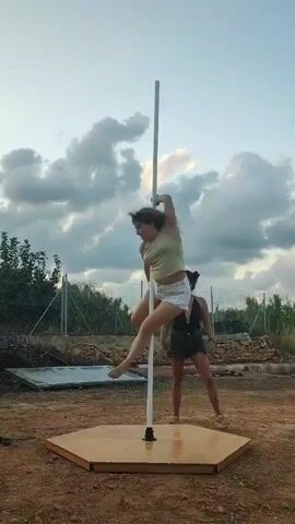 The video shows two women outdoors in a rural setting with a pole dancing setup. The woman on the pole is performing a routine, swinging and holding various poses. The second woman stands nearby, observing her. The pole is mounted on a hexagonal base on the ground. The background includes a fence, vegetation, and a cloudy sky.