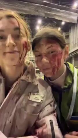 Two fair-skinned individuals, likely female, stand close together, posing for a selfie-style video. They are both wearing what appears to be stage makeup resembling blood and facial injuries. The person on the left wears a light-colored jumpsuit with a name tag reading "EMMA JOHNSON" partially visible. The jumpsuit is heavily splattered with what looks like blood. The person on the right wears a green high-visibility vest. They both have similar makeup patterns around their eyes and cheeks, simulating cuts and bruises. The background suggests they are on a film or theater set. There are metallic structures and scaffolding visible, giving the impression of a large, industrial-type space. The lighting is bright and diffuse. Initially, both individuals maintain a slight smile, then one begins to laugh.