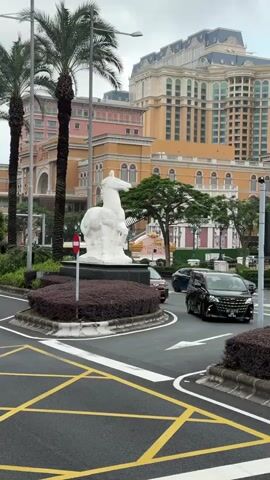 The video shows traffic circulating around a roundabout. At the center of the roundabout is a white horse statue atop a bed of shrubs. Several cars, taxis, and buses pass by in both directions. In the background, tall palm trees frame large, ornate buildings, with one building appearing particularly grand, featuring a tiered design and intricate detailing. The sky is overcast, suggesting a cloudy day. The road is marked with yellow lines, indicating traffic flow and safety zones.