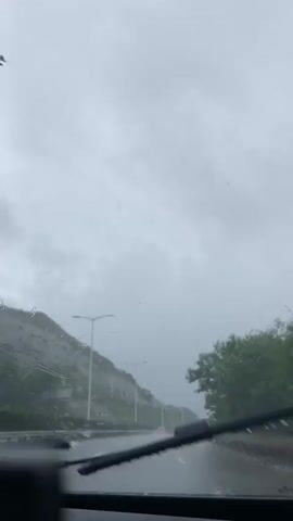 The video shows a view from inside a vehicle on a highway during rainfall. The road is wet and there are raindrops on the windshield. On the left side of the road, there is a hillside covered with vegetation and rocks, with a white barrier and streetlights running along the edge. On the right side, there are trees. Several vehicles are visible on the highway, including cars and a truck. The sky is overcast and appears grey. The overall atmosphere is moody due to the rainy weather.