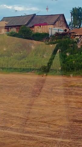 The video shows a rural landscape with a wooden house on a grassy hill. The sky is cloudy and the lighting suggests it is late afternoon or early evening. There are some small, blurry objects visible in the air, possibly insects or dust. A dirt road or clearing is in the foreground.