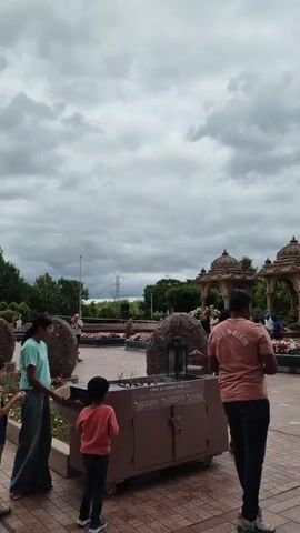 This video captures an outdoor scene at the BAPS Swaminarayan Akshardham temple in Robbinsville, New Jersey. The main subjects are people interacting within the temple grounds and a prominent golden statue.

In the foreground, several individuals are visible. A woman in a light blue t-shirt and jeans stands to the left, holding an object. Next to her, a young boy in a pink sweater is looking down. A man in a coral-colored t-shirt with "Calvin Klein" visible on the back stands facing away from the camera, looking towards the center of the frame. He appears to be holding something in his right hand. Further in the background, other people are seen walking and observing the surroundings.

The setting is a paved area with decorative tiles, surrounded by flower beds and large, natural-looking rocks. Behind the people and foreground elements, there are ornate structures with domes, characteristic of Indian temple architecture. In the distance, trees and a power line tower are visible. The sky is overcast with dramatic, heavy clouds, suggesting it might be a cloudy or potentially rainy day.

The activity appears to be a casual visit to the temple. People are walking, observing, and possibly participating in a ritual, as suggested by the man in the foreground holding an object that could be an offering. There is a sign on a brown structure in the foreground with text that appears to say, "Offer an Incense With Us."

A striking feature is the large golden statue of a man in a yoga pose, with arms raised towards the sky. This statue is positioned on a raised platform and is a focal point of the scene. The overall mood is serene and contemplative, typical of a spiritual site. The time of day is not explicitly indicated, but the cloudy sky suggests it could be daytime, possibly afternoon.