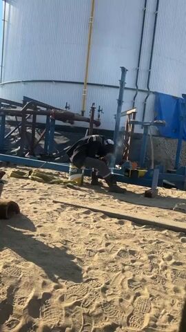 The video shows a person welding in a sandy outdoor industrial area. The person is wearing a dark jacket, dark pants, and a white hard hat. They are crouching down, bent over a piece of metal, and a bright light from the welding torch is visible, emitting smoke. The welding equipment is on the ground next to them. In the background, there is a large white cylindrical structure, possibly a storage tank, and a blue tarp. There are also pipes and metal beams present. The ground is covered in sand with visible footprints. The lighting suggests it is daytime, with shadows indicating the sun is casting light from the right.