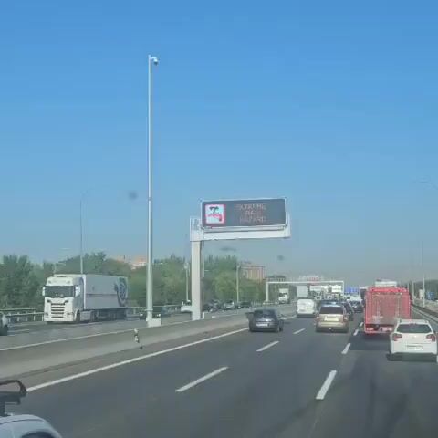 The video shows a highway scene with several vehicles. A digital sign mounted on an overhead gantry displays the text "EXTREME FIRE HAZARD" along with a graphic of a flame. Cars, trucks, and vans are driving on the multi-lane highway. The sky is clear and blue, and there are trees and buildings in the background. The camera appears to be mounted inside a vehicle, looking forward.