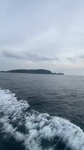 The video depicts a boat moving across a body of water under a cloudy sky. The sun is attempting to shine through the clouds, creating a glinting path on the water's surface. In the distance, a landmass, likely an island or coastline, is visible. The boat leaves a trail of white foam in its wake as it moves.