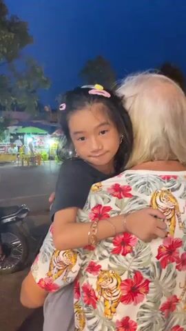 Here's a description of the video: 

The video shows a young Asian girl, possibly around 6-8 years old, being held by an elderly woman with short white hair. The girl is wearing a black t-shirt, has dark hair with pink and yellow hair clips, and is wearing bracelets. She's looking down, appearing a bit sad or pensive. The woman is wearing a brightly colored shirt with a pattern of tigers and red hibiscus flowers. She has her arms around the girl, holding her close. 

The background shows a street scene at dusk or night. There are street vendors and other people moving about. A motorcycle is parked nearby. The overall impression is one of comfort and affection between the child and the elderly woman.