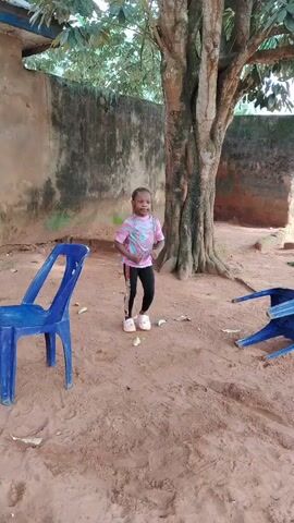 A young girl, approximately 4-7 years old, is energetically dancing outdoors on a sandy-brown dirt ground. She has dark hair styled in braids adorned with light-colored beads. She is wearing a multi-colored, possibly tie-dye, short-sleeved t-shirt (pink, blue, yellow, and white tones), black leggings, and light-colored sneakers. Her expression is joyful and lively as she moves her body.

The background features a weathered concrete wall, partially covered with moss or dirt, and a large tree with a thick, textured trunk and visible foliage. To the left, an upright blue plastic chair is visible, while another blue plastic chair lies overturned on the right side of the frame. The setting appears to be a compound or backyard in Akwa Etiti, Nigeria, under bright, natural daylight. The ground is unpaved, with some scattered dry leaves. A man's voice can be heard interjecting "Jesus" in the audio.