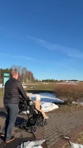 A man is pushing a stroller with a small child along a paved path near a body of water. Patches of snow are on the ground and the ice is melting around two multi-story buildings on a small island in the distance. The weather is sunny with a bright blue sky. The man is wearing a black jacket and the child is wearing a beige outfit with a hat.
