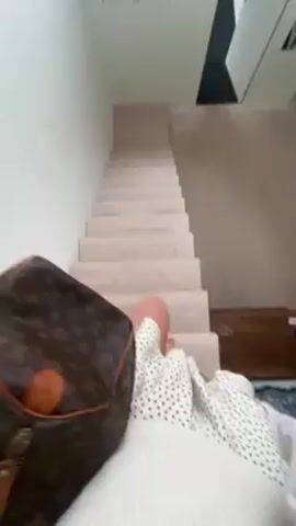 Here is a concise description of the video:

The video shows a first-person view of someone sitting on a staircase. The person is wearing a white sweater with black polka dots and a light-colored skirt. A Louis Vuitton bag is next to them. The staircase is light-colored wood. The walls are white, and there is a small black geometric design on the ceiling above the stairs. The video appears to be taken indoors.