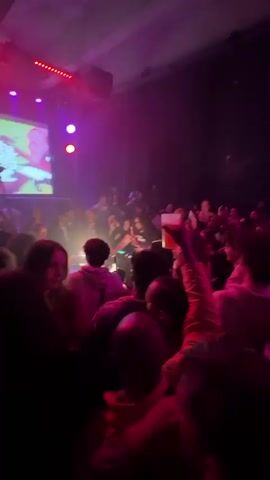 The video captures a vibrant scene from a crowded event with pink and purple lighting. The focus is on a DJ, a dark-haired woman positioned behind DJ equipment. The crowd is diverse, with individuals facing the DJ, some visible clearly while others are partially obscured. One person in the crowd holds up a drink. A screen displays graphics with the words "Ba Da Da DJ's" visible. The environment suggests a nightclub or party setting with dark curtains in the background. Red lights and a spotlight add to the dynamic atmosphere.