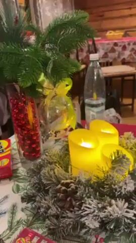 A video captures a festive indoor scene at what appears to be a New Year's celebration. The foreground displays a decorated table with candles and a Christmas arrangement. The camera pans back to reveal a room featuring a man with a bald head, a beard, wearing a orange sleeveless top, and green-striped leggings, sitting on a stool and playing a guitar. Another man is standing nearby, and a woman is seen in the background. The kitchen area with a refrigerator and wooden cabinets is visible behind the seated musician. The overall atmosphere suggests a cozy, intimate gathering.