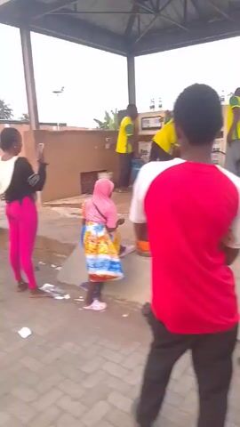 People are lined up at a gas station. The gas station is identified as DEE-TAWAK OIL & GAS by signage on the pumps. Three men in yellow and green uniforms are filling gas canisters for the customers. Some of the customers are standing while others are seated or squatting on the ground. One person is taking pictures. There are various colored canisters visible, including blue, orange, green, and brown. The area is paved with brick tiles. The sky appears overcast, and there are some buildings and vegetation visible in the background.