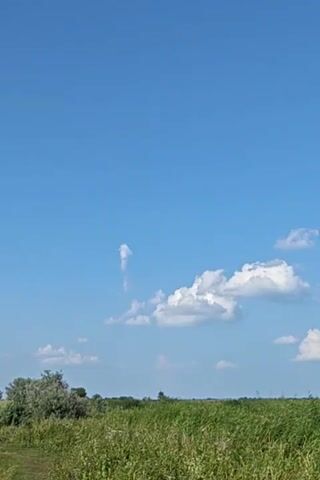 The video shows a bright blue sky with scattered white clouds. In the foreground, there is a field of tall green grass and some trees. A dirt path runs along the left side of the frame. The camera is static and focused on the landscape. There are no people or animals visible in the video. The scene appears to be a rural or natural setting on a clear day.