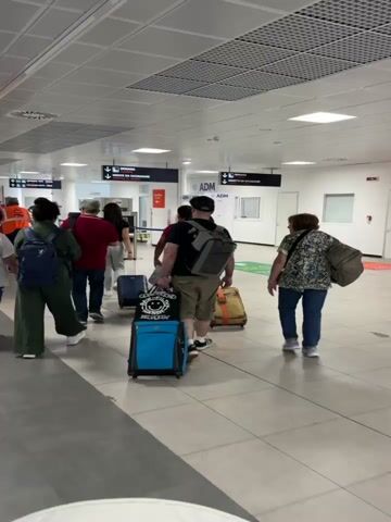 The video shows a group of people walking through an airport terminal. Most of the people are pulling luggage or carrying bags, heading toward customs, with signs indicating "Dogana," and "Niente Da Dichiarare." The terminal has a white tiled floor and a suspended ceiling with square lighting panels. The people are wearing a variety of casual clothing. The camera follows the group from behind.