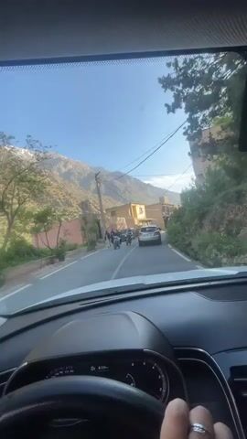 From the interior perspective of a vehicle, the camera captures a drive down a rural road, set against the backdrop of mountains. The road is bordered by buildings of varying heights and appearances. The driver's hand, adorned with a ring, is visible on the steering wheel. Ahead, a silver car travels in the same direction.  Pedestrians and individuals on bicycles can be seen on the side of the road. The sky is clear and blue.
