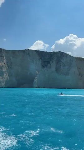 The video shows a scenic view from a boat on a bright, sunny day. The water is a vivid turquoise blue, with some white foam from the boat's wake. On the right side of the frame, a large, sheer cliff face rises dramatically from the sea. The cliff is a pale, rocky color with some green vegetation growing on top. In the distance, a small white boat with people on it can be seen near the base of the cliff. The sky is a clear blue with a few scattered white cumulus clouds. The camera appears to be moving forward, giving the impression of traveling along the coastline.