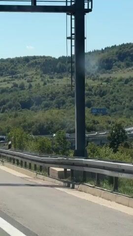 A bright sunny day with a clear blue sky and scattered white clouds. The video shows a highway with multiple lanes and overpasses, surrounded by lush green hills covered in dense forests and some open fields. In the foreground, a metal guardrail lines the side of the road. Cars and trucks are visible driving on the highway. A green highway sign with white lettering indicates "Mokro Polje 635 m." The overall impression is of a scenic, rural highway with active traffic.