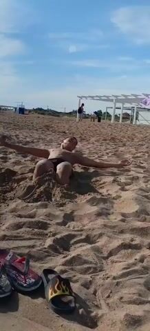 A shirtless boy in black swim trunks is standing on a sandy beach. He is partially buried in the sand up to his knees, with his arms outstretched. He smiles as he tries to free himself from the sand. The sky is bright blue with a few clouds. The sun shines brightly. In the background, other people are visible, and there are some white structures, possibly gazebos or beach huts. A pair of flip-flops is visible in the foreground, near the bottom left corner of the frame. The boy has dark hair and a light complexion. He looks like he is enjoying himself.