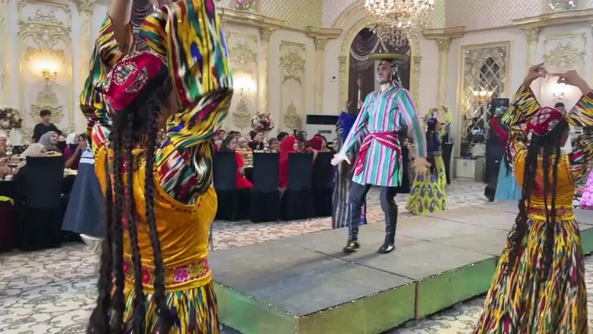 The video displays a vibrant cultural performance taking place in a grand indoor setting, likely a banquet hall, in Bekabad, Uzbekistan. The scene is characterized by opulent decor, including ornate walls with gold accents, large arched windows draped with luxurious curtains, and a prominent crystal chandelier hanging from the ceiling. The floor is covered in a patterned carpet.

The primary subjects are people engaged in a traditional dance. Several individuals, predominantly women, are dressed in colorful, intricately patterned traditional clothing. These outfits feature long, flowing dresses, often in yellow, with bright, geometric designs in various colors like red, blue, green, and gold. Many of the women have long, braided hair extensions, some adorned with decorative headwear. A male performer is also present, wearing a striped tunic with a sash and dark boots, also appearing to be part of the dance.

The activity is a cultural dance performance, indicated by the coordinated movements and the stage set up in the center of the room. The dancers are moving rhythmically, with some holding their arms aloft. The audience, seated at tables in the background, appears to be observing the performance. The atmosphere seems festive and celebratory, suggested by the elaborate attire and the engaging nature of the dance.

Notable details include the time of day being ambiguous due to the indoor lighting, but the overall ambiance suggests an evening event. The setting's grandeur points towards a significant cultural or social gathering. The clothing worn by the performers is a strong indicator of Uzbek or Central Asian cultural traditions.

There is some visible text on a black garment worn by a person in the left foreground, partially obscured, which appears to be a logo or text in white lettering, possibly related to an organization or event production. However, the text is not clearly legible.