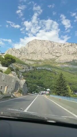 A car is driving on a road with a mountain in the background. The sky is blue with white clouds. There are trees on the side of the road and a speed limit sign that says 40.
