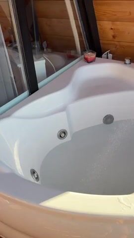 A corner bathtub is filling with water. The bathtub is white and has several jets. There are some knobs and a faucet visible. A red candle is also on the edge of the tub. The tub is situated on a wooden deck, and beyond the deck is a view of a lake, houses, and trees. The sun is shining, indicating it is daytime. The video is shot from a first-person perspective.