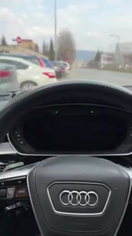 The video shows the interior of a car, specifically an Audi, from the driver's perspective. The focus is on the dashboard, steering wheel, and the road ahead. The driver is stationary initially, then begins driving down a street. The landscape outside includes buildings, trees, parked cars, and street lights. The car's infotainment system is visible, displaying radio information. An air freshener hangs from the rearview mirror. The overall environment appears to be a relatively quiet suburban area on a cloudy day.