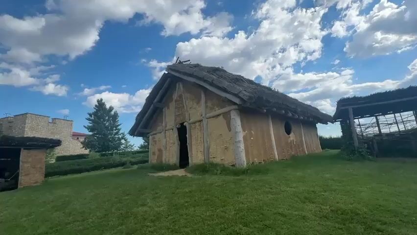 The video pans across a grassy area with several small, primitive-looking buildings constructed from mud and straw with thatched roofs. A pathway of dirt and gravel winds through the scene. In the background, a stone fortress-like structure stands against a bright blue sky dotted with white clouds. Trees are scattered throughout the landscape, and some of the buildings appear to be part of an historical or archaeological exhibit. The overall impression is of an outdoor historical reconstruction.