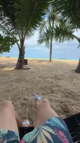 A person lies on a beach, viewed from a first-person perspective, with their legs and feet visible in the foreground. They are wearing floral-patterned swim trunks and light blue flip-flops with a white design. Their legs are extended towards a sandy beach dotted with palm trees. In the background, the ocean meets the horizon under a cloudy sky.