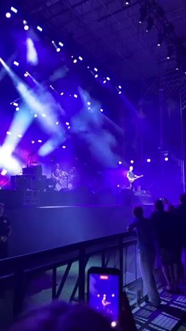 The video shows a concert stage from a slightly elevated perspective behind a railing.  In the foreground, the hands of a person are visible holding a smartphone, recording the event, with a blurry image of the stage on the phone screen.  Behind this person, several other audience members are standing, looking towards the stage.  The stage itself is brightly lit with purple and white spotlights, with smoke effects filling the air.  Several musicians are performing on stage.  One musician is clearly visible in the center right, playing a guitar and singing. To the left, another musician appears to be playing drums.  The overall atmosphere is that of a live musical performance with dramatic lighting.