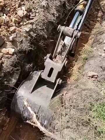 The video shows an excavator bucket digging into a trench. The excavator arm is extended and the bucket is scooping dirt and rocks. The trench is narrow and appears to be dug into a hillside with loose soil and debris. The surrounding environment is grassy with some scattered rocks.