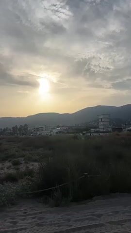 The video begins with a landscape shot of a beach with mountains in the background and a cloudy sky. The sun is visible through the clouds. The camera then pans across the beach, showing sand dunes and a fence line. The next shot is a close-up of a white paper bag with a logo that says "COMPA". The video ends with a shot of feet walking on a wooden boardwalk next to the sand.