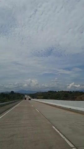 A vertical, wide shot shows a highway with light traffic under a partly cloudy sky. The highway stretches into the distance with trees and hills on either side. The road is concrete with white lane markers. Two vehicles are visible in the distance, driving away from the camera. The sky is filled with scattered white clouds against a blue backdrop. The overall impression is of a journey on a clear day.