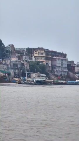 The video shows a wide shot of a river with buildings lining the embankment. Several boats are visible on the water, and there are many structures built on the hillside. The sky is overcast. The buildings are multi-story and appear to be of various ages and architectural styles. Some are residential, while others may be commercial or religious. The boats are of different sizes and types, suggesting various uses such as transportation or tourism. The overall impression is of a busy urban waterfront.