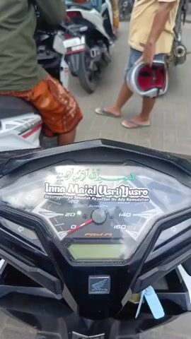 The video shows a gas station with several people and motorcycles. In the foreground, the dashboard of a motorcycle is visible with Arabic text and the words "Inna ma'al 'usri yusra" on a sticker, which translates to "Verily with hardship there is relief." A person wearing a green shirt and orange shorts is seated on the motorcycle. In the background, a man in a tan shirt and denim shorts is holding a red and white helmet and standing next to a motorcycle. Other motorcycles and people are seen at the gas pumps. A green truck is also present. The scene appears to be a busy gas station.