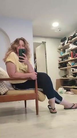 The video begins with a woman sitting in a wooden chair, holding a smartphone and taking a selfie. She has long, curly blonde hair and is wearing a yellow t-shirt and black leggings with white stripes. Her left arm is resting on the armrest of the chair. Her right leg is bent at the knee and her right foot is resting on a pillow. Her left leg is extended and her left foot is on the floor. She is wearing black flip-flops. Behind her, to the right, is a shelf unit filled with shoes and other items. To the left of the woman is a doorway and a wall. The video then cuts to a shot of a tripod with a camera attached. The tripod is set up in front of a table with various equipment on it, including a fan and what appears to be a lighting setup. The background is blurred, but it seems to be an indoor space with shelves filled with items on the left side. The video then returns to the woman taking the selfie. The camera angle shifts slightly, showing more of the room behind her, including a shelf filled with many pairs of shoes and other items. The lighting in the room is bright, with a ceiling light visible.