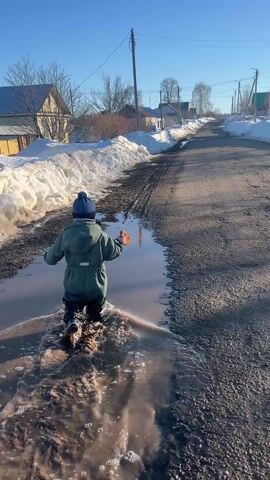 A small child dressed in a green snowsuit and blue knit hat is seen from behind, splashing through a large, muddy puddle on a paved road. The road is wet and lined with patches of snow on either side. Houses are visible in the background, along with trees bare of leaves. The sky is bright blue. The child holds a small orange object in their hand as they walk through the puddle, creating splashes. The road slopes uphill into the distance. The overall scene suggests a rural or suburban area in early spring, with melting snow creating muddy conditions.