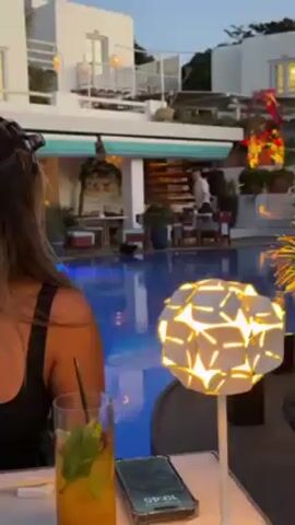 Here's a brief description of the video:

The video is shot from the perspective of someone sitting at a table outdoors near a pool. The scene is a restaurant or resort patio area, with white buildings in the background.  A woman with long hair is partially visible in the foreground. On the table, there is a drink, a phone showing 19:45, and a decorative lamp.

The focus of the video quickly shifts to a small dark animal, possibly a cat, that walks along the edge of the pool. The animal appears to be sniffing around. The general environment is calm and well-lit.
