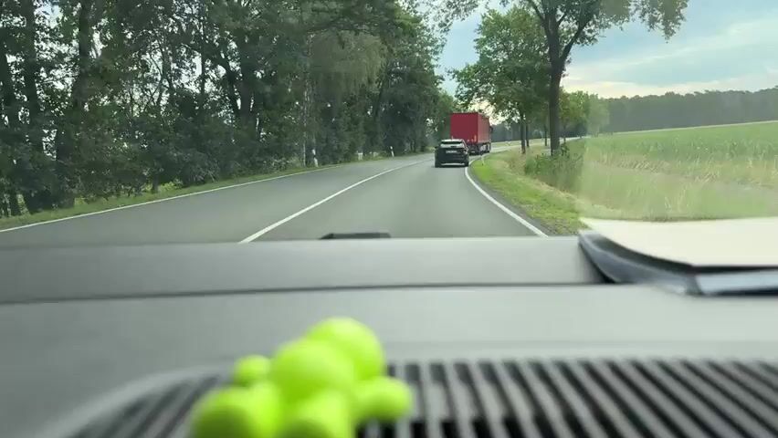 The video shows a view from inside a car driving on a road lined with trees. In the foreground, a blurred green object, possibly a toy, is visible on the dashboard. Ahead on the road, a dark-colored car is following a red truck. The sky is overcast. There are fields visible on the right side of the road.