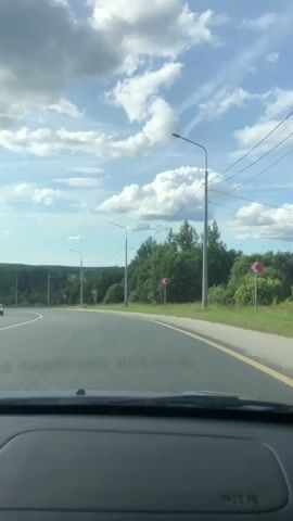 This is a first-person perspective video taken from inside a car looking out at a curved road on a cloudy day. The road has a white line on the left and a yellow line on the right. There are streetlights and utility poles lining the right side of the road. Lush green trees and foliage line the roadside and form a distant landscape. There are no people visible in the video. The car is in motion.