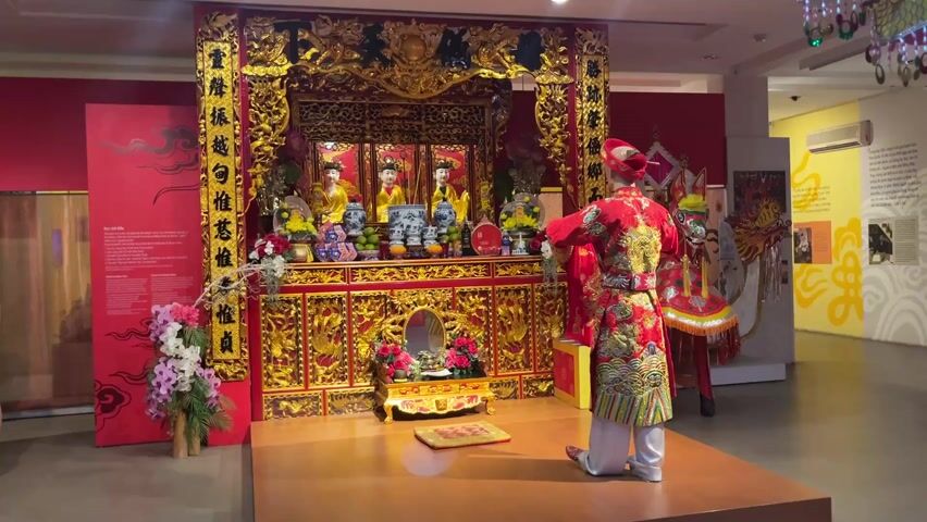 The video captures a person dressed in a traditional red and gold Vietnamese outfit, standing on a wooden platform in front of an elaborate, gilded altar. The altar is adorned with statues, bowls of fruit, flowers, and a small mirror. Intricate carvings and decorations cover the altar. The room appears to be a museum or exhibit space, with red-painted walls, informational signs, and decorative elements. Other people are visible in the background, moving around the room and observing the exhibit. The overall scene suggests a cultural or religious display, possibly reenacting a traditional ceremony or ritual.
