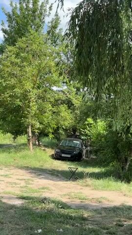 A black car is parked on a dirt path surrounded by trees and grass. A black folding chair is placed in front of the car. The camera pans to the right, revealing a body of water with reeds and more trees in the background. The sky is visible, with some clouds.