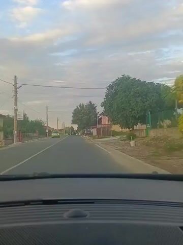 The video captures a point-of-view perspective from inside a vehicle as it travels down a paved road. The road appears to be a two-lane street in a rural or suburban area, lined with houses and trees on either side. The sky is partly cloudy, with daylight suggesting it is either morning or late afternoon.

On the left side of the road, there is a sidewalk, a fence, and houses. A few trees are visible, and a utility pole with wires runs along the side of the road. A bright green car is seen traveling in the opposite direction on the left lane.

On the right side of the road, there are more houses with fences. A large tree stands prominently near the roadside, with its branches extending over the road. Further down the road, a dark-colored car is parked on the right shoulder, and then a white SUV with a spare tire mounted on the back is also parked on the right side. The road ahead curves slightly to the left. The dashboard of the vehicle from which the video is recorded is visible at the bottom of the frame.